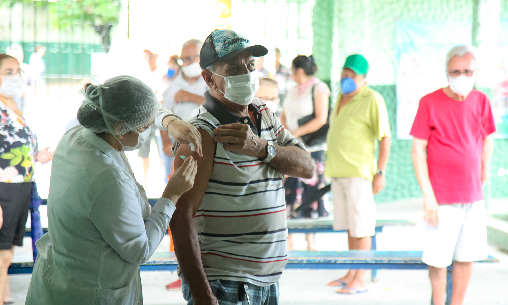 enfermeira vacinando senhor de máscara e outros idosos ao fundo aguardando